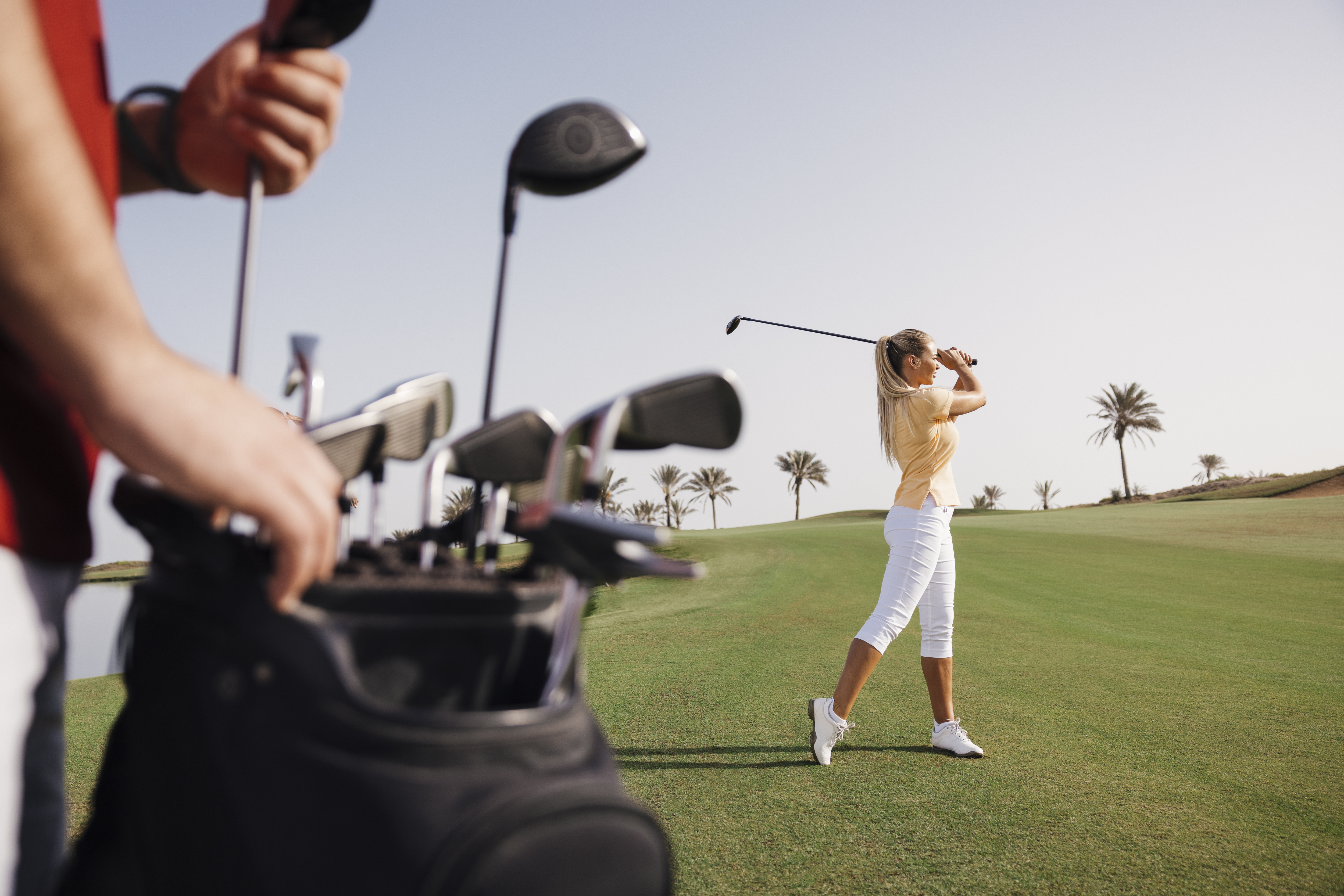 A golfer swinging a golf club at Saadiyat Beach Golf Club, with golf clubs in the foreground and palm trees in the background.